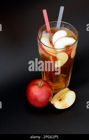 Ein großes Glas facettierten Fruchtsaft mit Eis, Obststücken und Strohhalmen auf schwarzem Hintergrund, daneben befindet sich ein reifer roter Apfel. Nahaufnahme. Stockfoto
