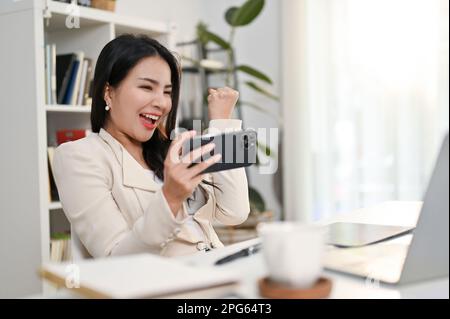 Fröhliche und aufgeregte asiatische Millennials-Geschäftsfrau oder Chefin, die auf ihren Telefonbildschirm schaut, ihren erfolgreichen Sieg feiert, Projekt genehmigt, zurückweisen Stockfoto