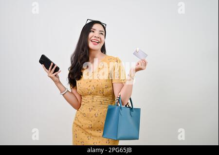 Lächelnde junge asiatische Frau in lässiger Kleidung mit Kreditkarte und Smartphone, während sie vor einem isolierten weißen Hintergrund steht. Online-Shopping Stockfoto