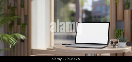 Nahaufnahme eines tragbaren Laptops mit weißem Bildschirm, Kaffeetasse und Dekorationspflanze auf einem minimalistischen Holztisch in einem schönen, modernen Café oder in Zusammenarbeit Stockfoto