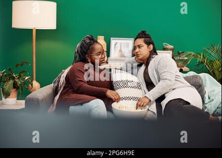 Die Wochenenden sind für Frauenübernachtungen gemacht. Zwei junge Frauen, die Popcorn trinken und sich zu Hause auf dem Sofa unterhalten. Stockfoto