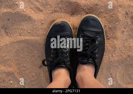 Weibliche Beine in schwarzen Sneakern stehen im Sommer auf dem Sand mit Blick von oben Stockfoto