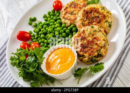 Krabbenkuchen mit Remouladesauce, grünen Erbsen, Tomaten, frischem Koriander auf weißem Teller auf weißem Holztisch, Nahaufnahme Stockfoto