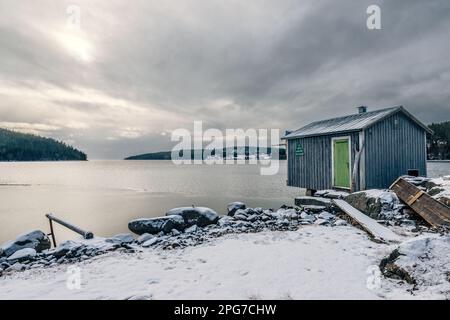 Eine Winterszene eines kleinen hölzernen Bootshauses an einer schneebedeckten Küste neben einem ruhigen See Stockfoto