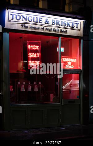 Ein Salt Beef Shop namens Tongue and Brisket, der nachts in Wardour Street, London, aufgenommen wurde, mit roten Neonschildern mit der Aufschrift Hot Salt Beef and Seating at Rear. Stockfoto