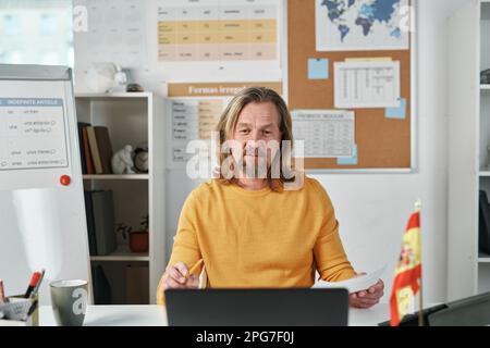 Reife Lehrerin sitzt am Tisch vor dem Laptop und spricht online im Unterricht mit Schülern Stockfoto