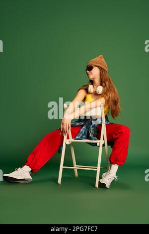 Ganzkörperfoto eines jungen Mädchens mit Kopfhörern, Sonnenbrille, auf einem Stuhl sitzend und mit grünem Hintergrund posiert Stockfoto