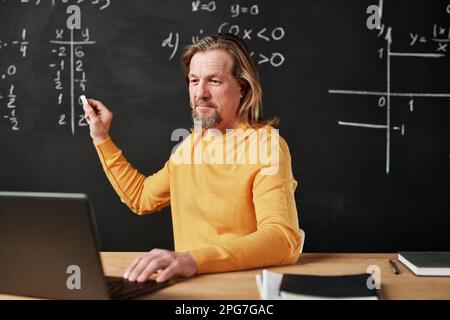 Reife bärtige Lehrerin, die an eine Tafel schreibt und den Laptop für den Online-Unterricht im Klassenzimmer verwendet Stockfoto