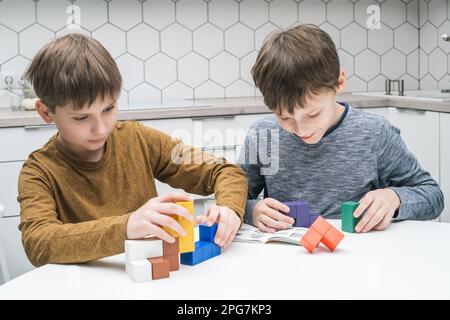 Lächelnde, konzentrierte kleine Jungen, die mit bunten Steinen spielen, sich miteinander verbinden und Figuren auf einem weißen Tisch bauen. Kinder lesen Regeln und Spiel Stockfoto