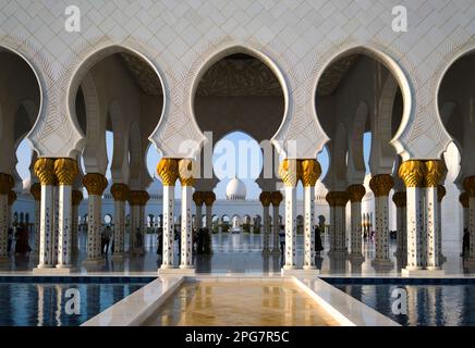 Blick auf den schattigen Wandportiko mit Säulen mit goldenen Palmenstämmen. In der Scheich-Zayid-Moschee in Abu Dhabi, Vereinigte Arabische Emirate, Stockfoto