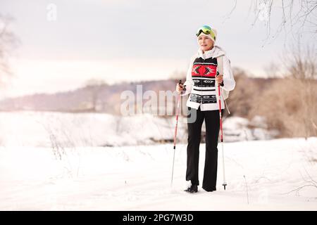 Glückliche Seniorin geht mit nordic Walking Stöcken auf weißem Hintergrund von Schnee und Natur. Sportlicher, aktiver Winterlebensstil Stockfoto