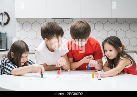 Vier fröhliche, lächelnde Kinder, die Brettspiele spielen und farbenfrohe Figuren auf dem Tisch in der Küche auswählen. Kinderentwicklung und Lernspiele und Spaß Stockfoto