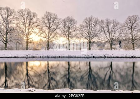 Eine ruhige Winterszene mit einem ruhigen Teich, eingebettet in einen Park, umgeben von schneebedeckten Bäumen, mit Sonnenuntergang im Hintergrund Stockfoto