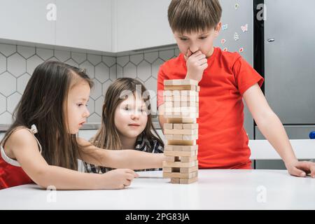 Drei Kinder mit konzentrierten, aufmerksamen Mädchen und einem Jungen, die auf dem Tisch ein Backsteinturmspiel spielen. Balanciere die Herausforderung und bewege Blöcke aus dem Turm Stockfoto