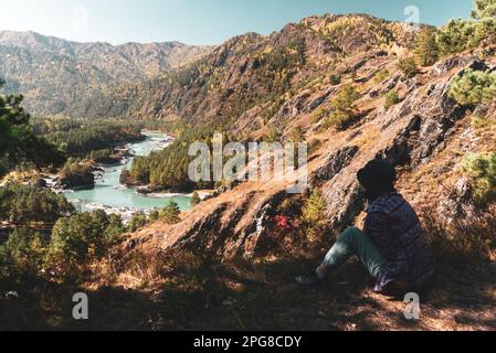 Ein Touristenmädchen sitzt tagsüber in Sibirien im Herbst neben einer Klippe vor dem Hintergrund eines Panoramas auf die Berge und den Fluss Katun in Altai. Stockfoto