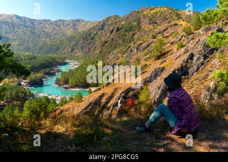 Eine Reisende sitzt tagsüber in Sibirien im Herbst in der Nähe einer Klippe vor dem Hintergrund eines Panoramas auf die Berge und den Fluss Katun in Altai. Stockfoto