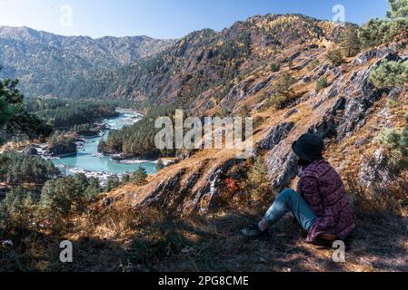 Eine Reisende sitzt tagsüber in Sibirien in der Nähe einer Klippe vor dem Hintergrund eines Panoramas auf die Berge und den Fluss Katun in Altai. Stockfoto