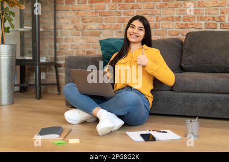 Glückliche spanierin, die Daumen-hoch-Geste zeigte und den Laptop benutzte, Hausaufgaben machte, eine Online-Schule empfahl Stockfoto