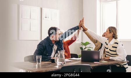 Erfolgreiche Geschäftsleute, die während eines Meetings in einem Büro lächeln und sich gegenseitig High Five geben. Eine Gruppe glücklicher Designer feiert ihren Sieg als a t Stockfoto