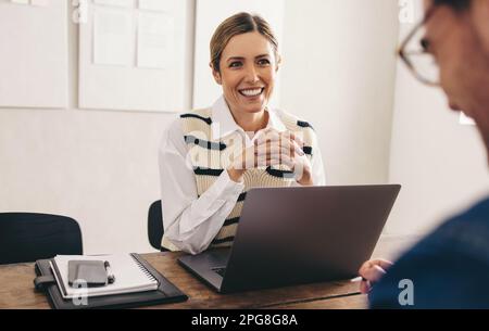 Weibliche einstellende Managerin, die glücklich lächelt, während sie einen Kandidaten in ihrem Büro interviewt. Eine fröhliche Geschäftsfrau, die ein Meeting mit einer App in der engeren Auswahl hat Stockfoto