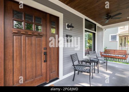 Eine hölzerne Eingangstür auf einer grauen überdachten Veranda mit Stühlen und einer hölzernen Hängeschwange. Stockfoto