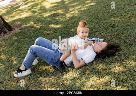 Brünette Mutter schmollt Lippen und liegt auf dem Gras mit einem kleinen Mädchen, Stock Image Stockfoto