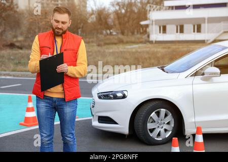 Kursleiter mit Klemmbrett in der Nähe des Fahrzeugs auf der Teststrecke. Fahrschule Stockfoto