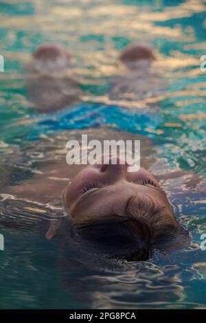 Der Junge liegt in türkisfarbenem Wasser mit geschlossenen Augen auf dem Rücken. Ein Junge, der sich im Sommer im Hotelschwimmbad erholt Stockfoto