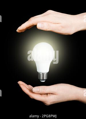 Leuchtende LED-Glühbirne in den Händen einer Frau auf schwarzem Hintergrund, Energieeinsparung, Weltwirtschaftskonzept, Stromprobleme, Stromausfälle aufgrund von Déstru Stockfoto