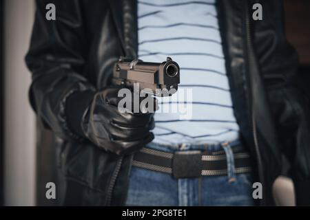 Gib mir dein Geld. Räuber zielt mit Pistole auf das Opfer. Ein bewaffneter Dieb mit Kapuze in Lederjacke hält die Waffe in der Hand. Waffe, Verbrechen. Stockfoto