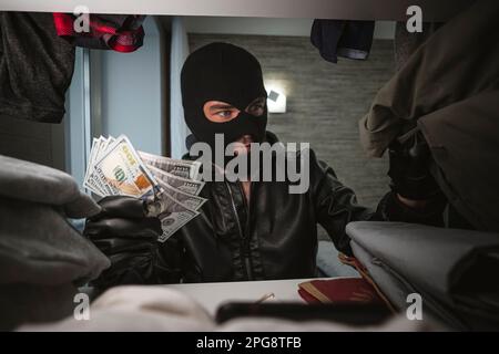 Ein Dieb in einer schwarzen Maske durchsucht einen Kleiderschrank auf der Suche nach Geld und Wertsachen. Konzept des Wohnungsdiebstahls. Kleidung stehlen. Stockfoto