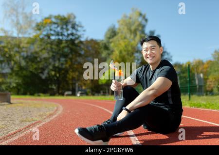 Junger asiatischer Sportler, der auf dem Laufband im Stadion sitzt und sich nach dem Joggen und Sport ausruht. Er hält eine Flasche, trinkt Wasser, erfrischt sich, löscht seinen Durst. Ein Lächeln schaut in die Kamera. Stockfoto