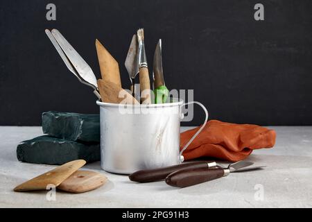 Werkzeuge zum Formen und Modellieren in einem Aluminiumbecher und einem Stück Plastik, Ton auf dunklem Werkstatthintergrund Stockfoto