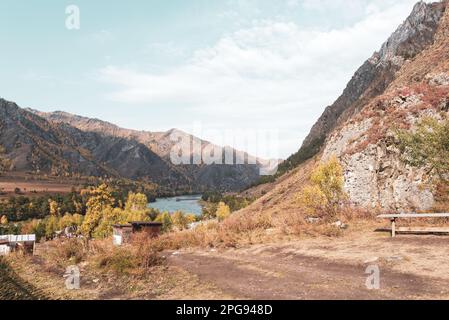 Eine Bank und Ferienhäuser auf einem Hügel inmitten der Berge in der Nähe des Flusses in Altai in Sibirien während des Herbsttages. Stockfoto