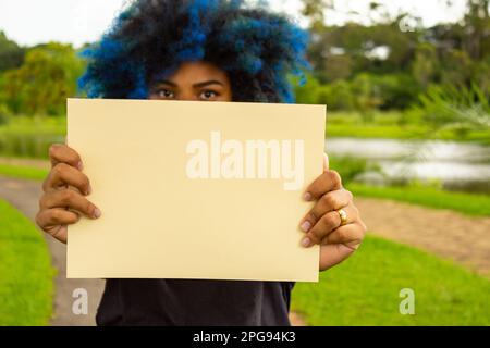 Goiania, Goias, Brasilien – 21. März 2023: Eine junge Frau mit blau gefärbtem Haar, hinter einem leeren Poster verborgenem Gesicht und einer Landschaft im Hintergrund Stockfoto