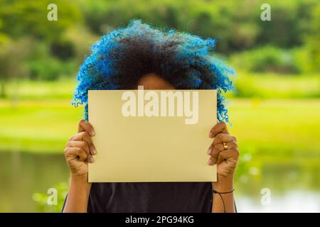 Goiania, Goias, Brasilien – 21. März 2023: Eine junge Frau mit blau gefärbtem Haar, hinter einem leeren Poster verborgenem Gesicht und einer Landschaft im Hintergrund Stockfoto