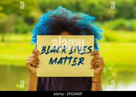 Goiania, Goias, Brasilien – 18. März 2023: Eine junge Frau mit blau gefärbtem Haar, deren Gesicht hinter einem Schild mit dem Text "Black Lives Matter" verborgen ist. Stockfoto