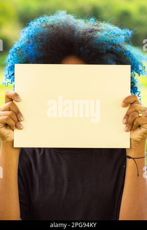 Goiania, Goias, Brasilien – 21. März 2023: Eine junge Frau mit blau gefärbtem Haar, hinter einem leeren Poster verborgenem Gesicht und einer Landschaft im Hintergrund Stockfoto