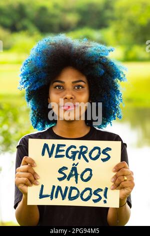 Goiania, Goias, Brasilien – 21. März 2023: Eine junge Frau mit gefärbtem blauem Haar, die ein Schild mit dem Text "Schwarze sind wunderschön" in der Hand hält. Stockfoto