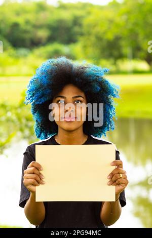 Goiania, Goias, Brasilien – 21. März 2023: Eine junge Frau mit gefärbtem blauem Haar, mit einem leeren Schild und einer Landschaft im Hintergrund. Stockfoto