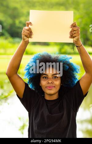 Goiania, Goias, Brasilien – 21. März 2023: Eine junge Frau mit gefärbtem blauem Haar, mit einem leeren Schild und einer Landschaft im Hintergrund. Stockfoto