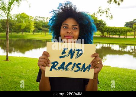 Goiania, Goias, Brasilien – 21. März 2023: Eine junge Frau mit gefärbtem blauem Haar, die ein Schild mit dem Text "Ende des Rassismus" in der Hand hält. Stockfoto