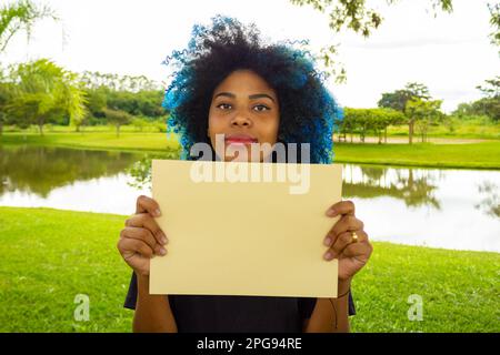 Goiania, Goias, Brasilien – 21. März 2023: Eine junge Frau mit gefärbtem blauem Haar, mit einem leeren Schild und einer Landschaft im Hintergrund. Stockfoto