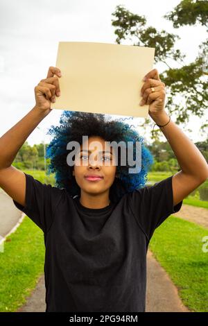 Goiania, Goias, Brasilien – 21. März 2023: Eine junge Frau mit gefärbtem blauem Haar, mit einem leeren Schild und einer Landschaft im Hintergrund. Stockfoto