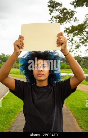 Goiania, Goias, Brasilien – 21. März 2023: Eine junge Frau mit gefärbtem blauem Haar, mit einem leeren Schild und einer Landschaft im Hintergrund. Stockfoto