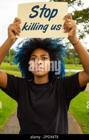 Goiania, Goias, Brasilien – 21. März 2023: Eine junge Frau mit gefärbtem blauem Haar, die ein Schild mit dem Text "Hör auf, uns zu töten" in der Hand hält, mit einer Landschaft im Stockfoto