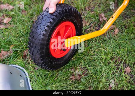 Auf der Straße ist eine Schubkarre aus Metall, ein Mann dreht eine neue Schubkarre für die Arbeit, zieht Schrauben und Muttern fest, hängt ein Rad auf Stockfoto