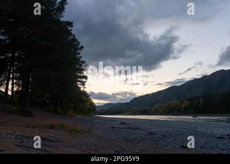 Die Sonne geht am Morgen in den Wolken über einem Berg in der Nähe des Flusses Katun an einem Steinstrand mit einem Wald in Altai in Russland auf. Stockfoto