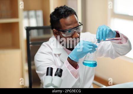 Junger schwarzer Mann in Arbeitskleidung, der Flüssigkeit aus dem Kolben in den Becher gießt, während er im wissenschaftlichen Experiment am Arbeitsplatz im Labor sitzt Stockfoto
