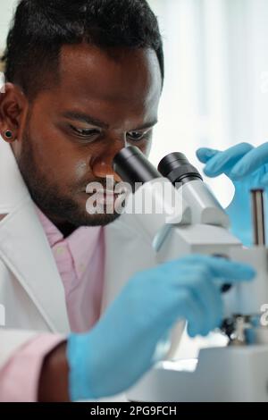 Junger männlicher Mikrobiologe in Schutzhandschuhen, der neue Viren oder Substanzen im Mikroskop während der wissenschaftlichen Laborforschung untersucht Stockfoto